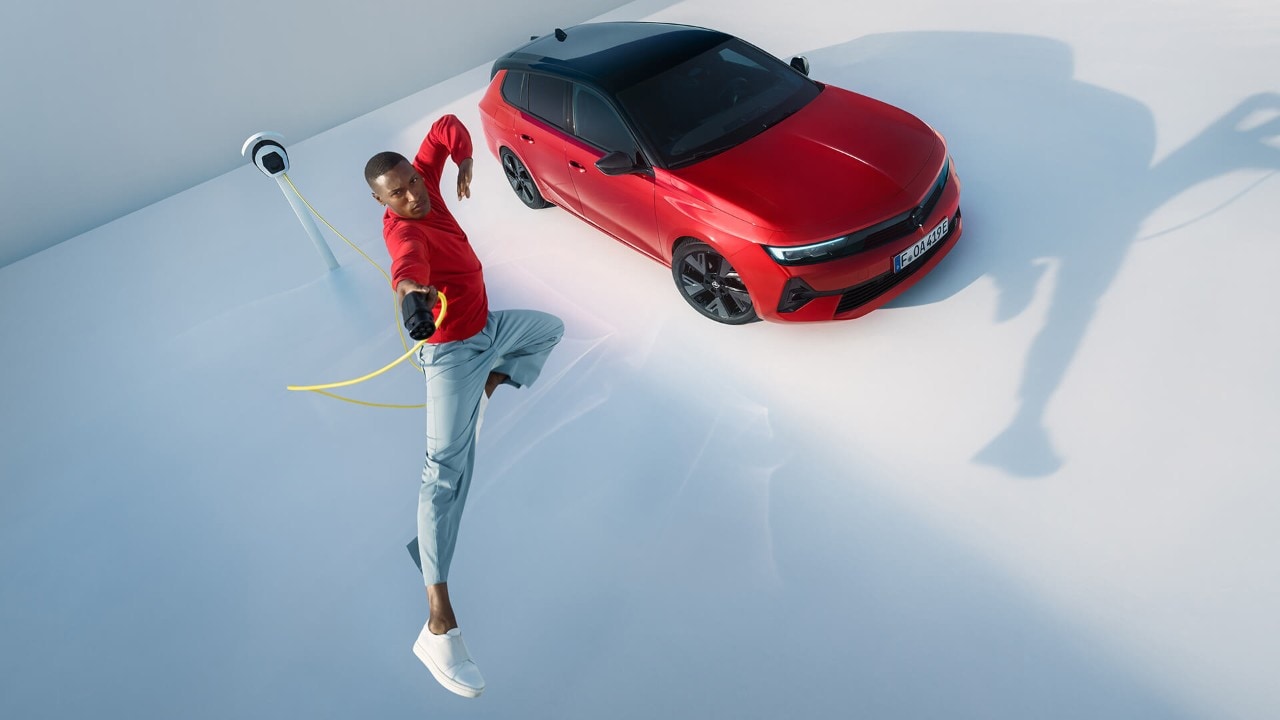 Muž skáče do vzduchu s nabíjacou stanicou pre elektrické vozidlá, v pozadí je Opel Astra Sports Tourer Electric