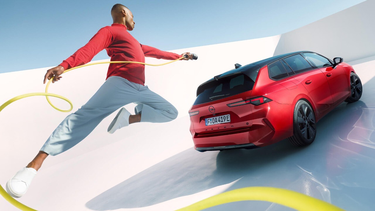 Muž skáče do vzduchu s nabíjacím káblom pre elektrické vozidlá, v pozadí je Opel Astra Sports Tourer Electric