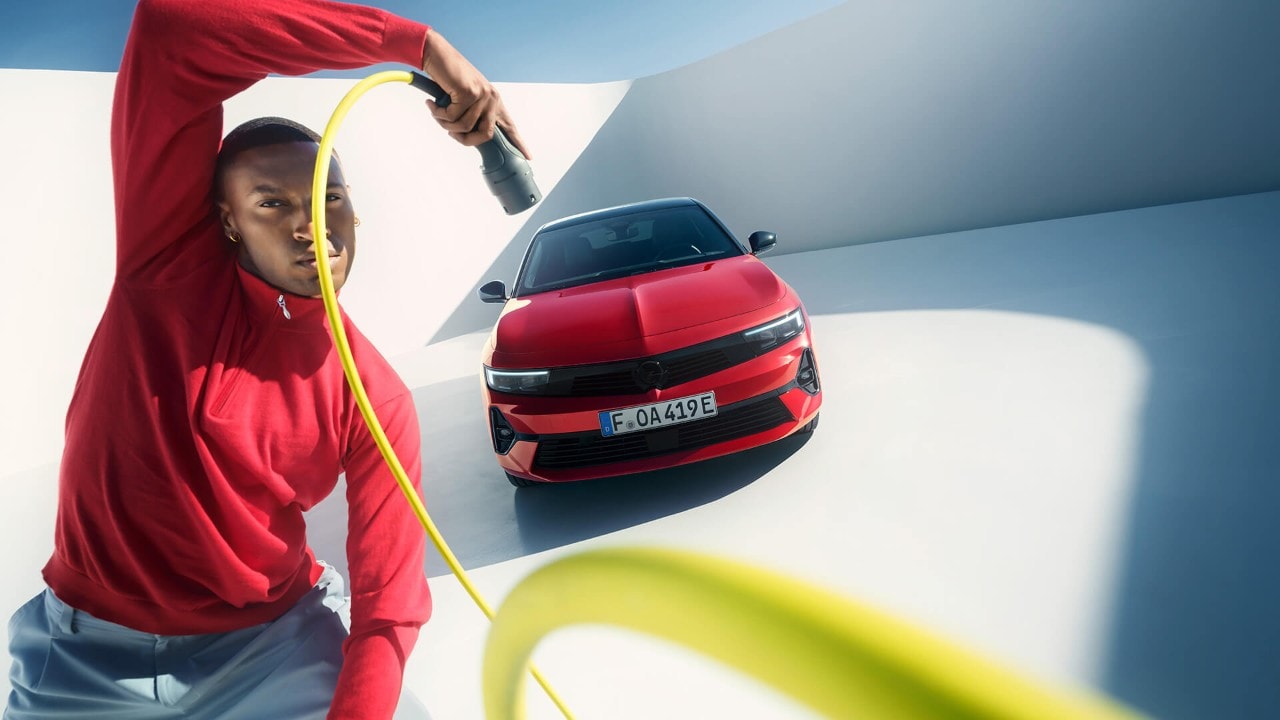 Muž stojí pred červeným modelom Opel Astra Sports Tourer s čiernou strechou a v ruke drží nabíjací kábel pre elektrické vozidlá